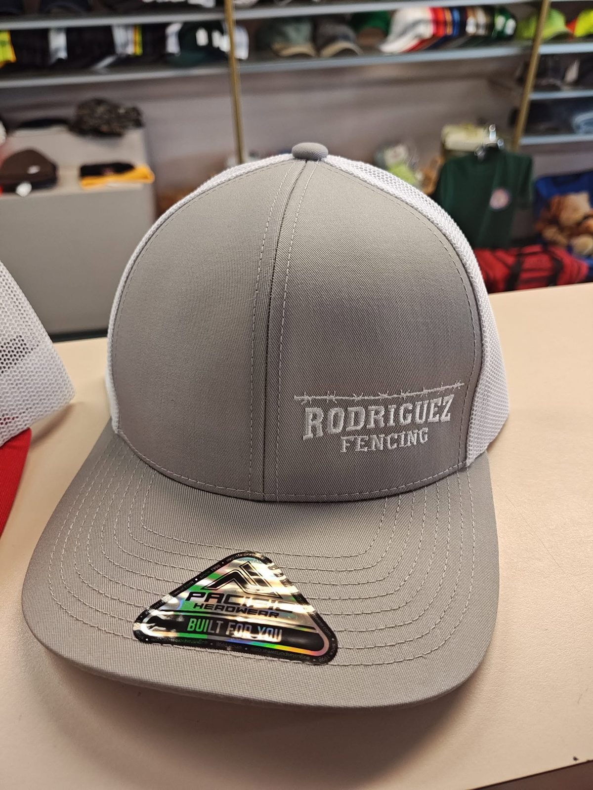 Grey and white mesh trucker cap embroidered in white with Rodriguez Fencing under a barbed wire line